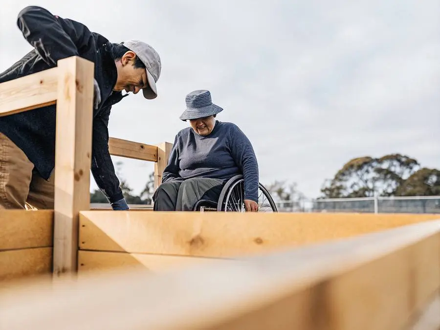 A man helping a woman in a wheelchair at a raised timber garden bed outdoors