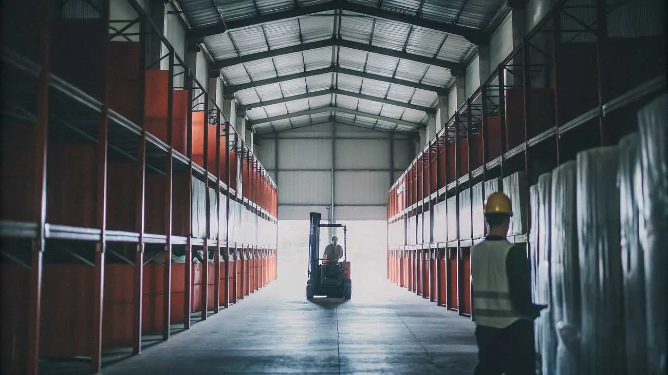 A forklift driving down a warehouse aisle, with a worker in a hard hat and hi-vis vest in the foreground